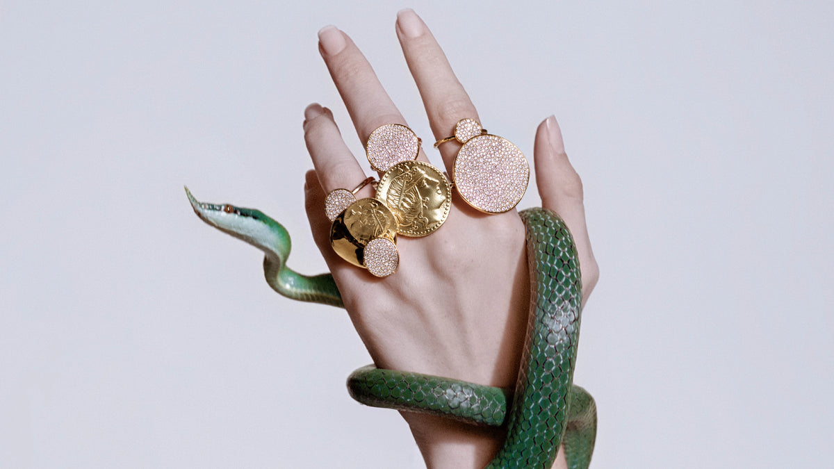 A hand with multiple gold and rose gold coin-style rings wrapped in a green snake, posed against a light gray background.