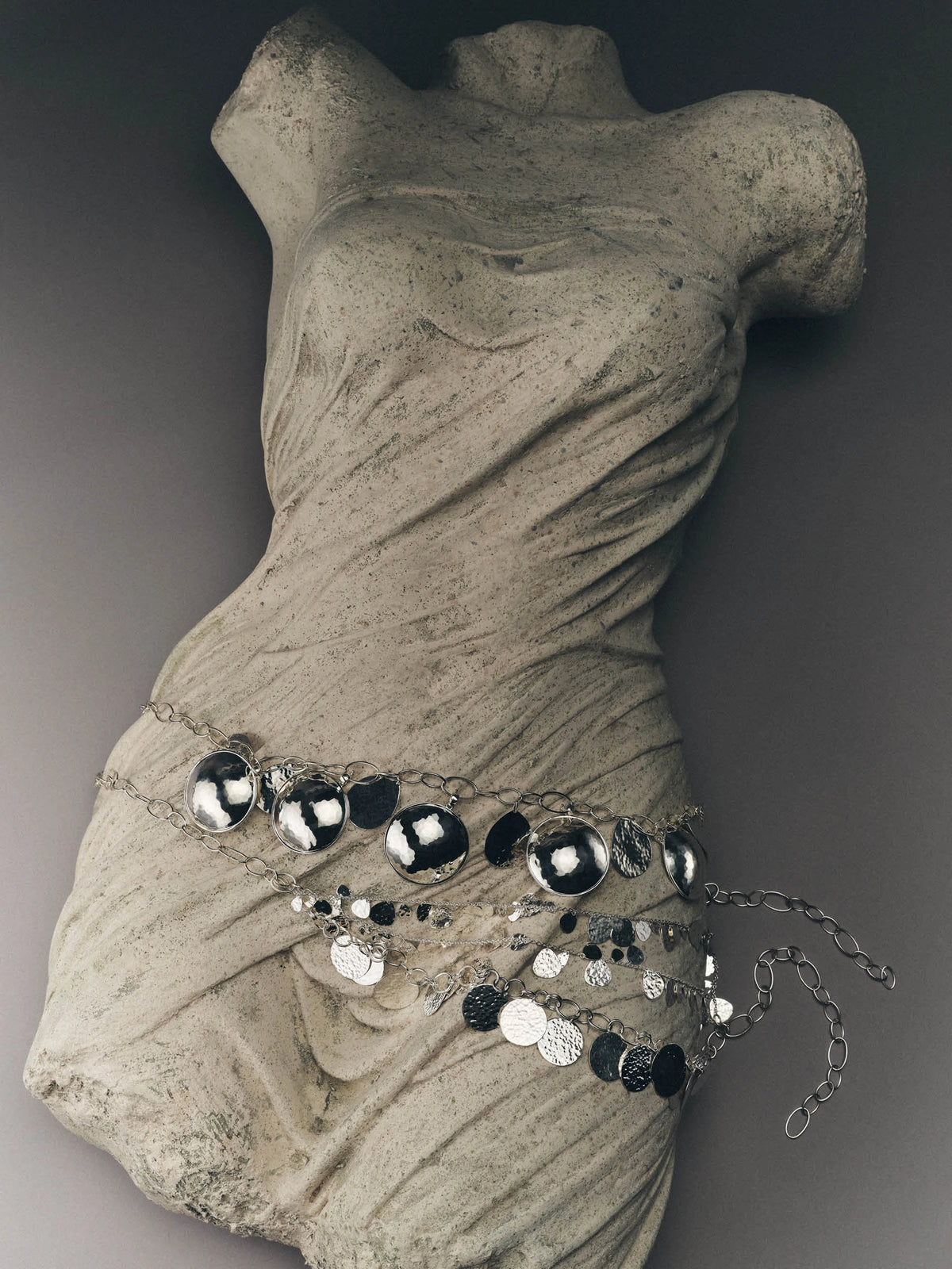 A silver-toned chain belt with large round black and silver discs and dangling coin charms, displayed on a draped stone torso mannequin against a dark gray background.