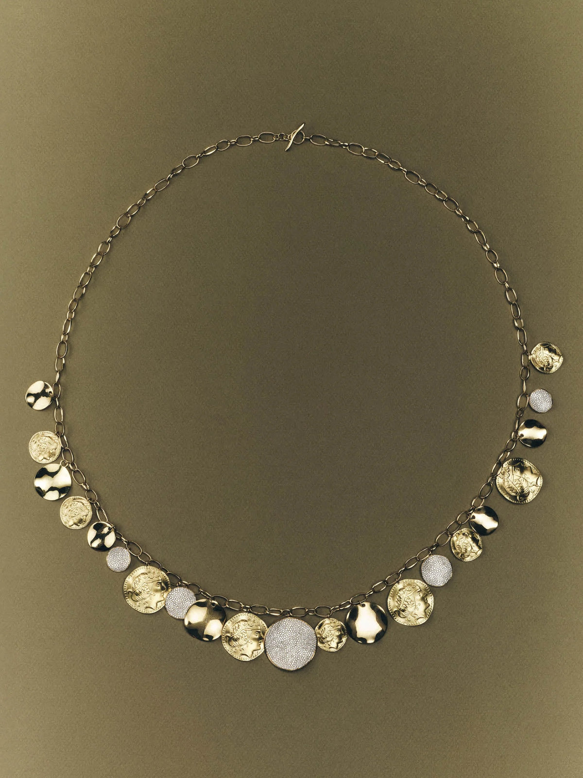 A statement necklace featuring a dark metal chain adorned with a series of dangling round and faceted discs in metallic gold, silver, and white, displayed against a solid dark olive background.